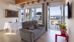 Ein Wohnzimmer mit einer Couch, einem Esstisch mit Stühlen, einem Gemälde und Meerblick durch offene Türen.