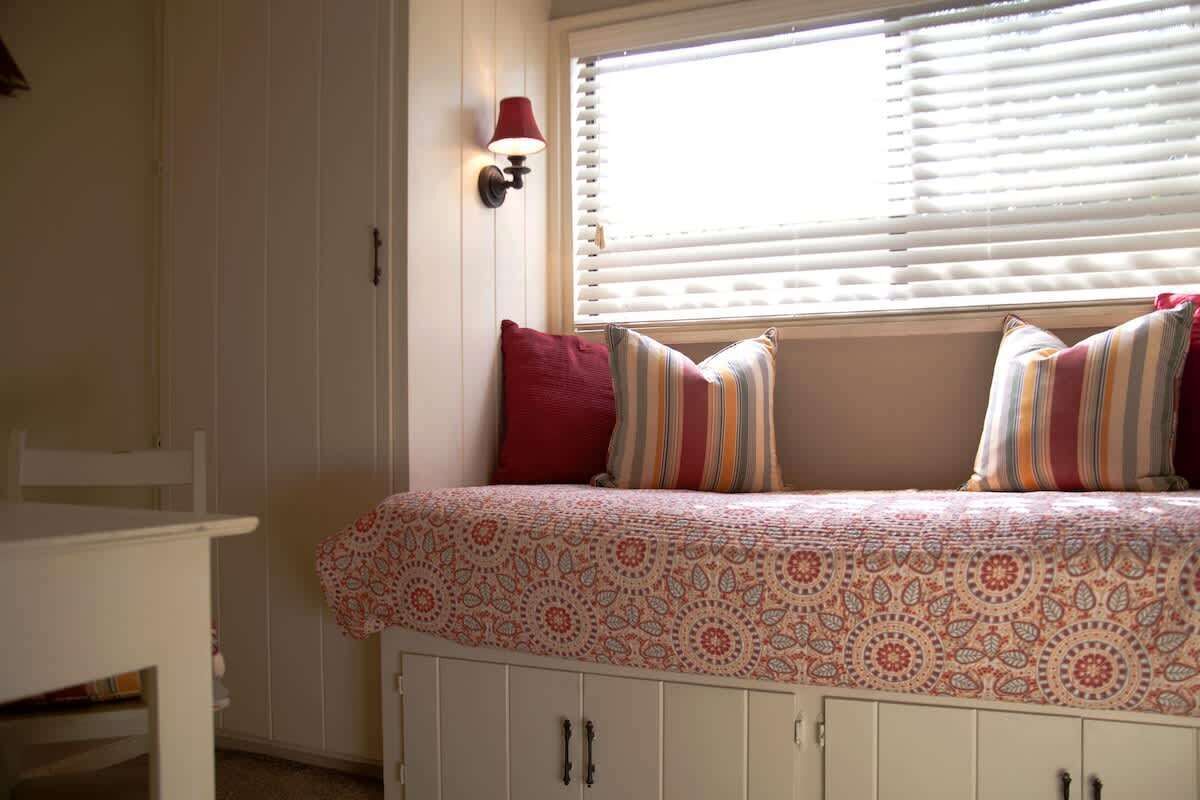 A bedroom with a bed, a window with blinds, a wall-mounted lamp, and a white chair.