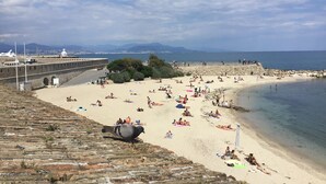 Una playa con gente tomando el sol y nadando, un muro de piedra y una vista del mar y montañas a lo lejos.