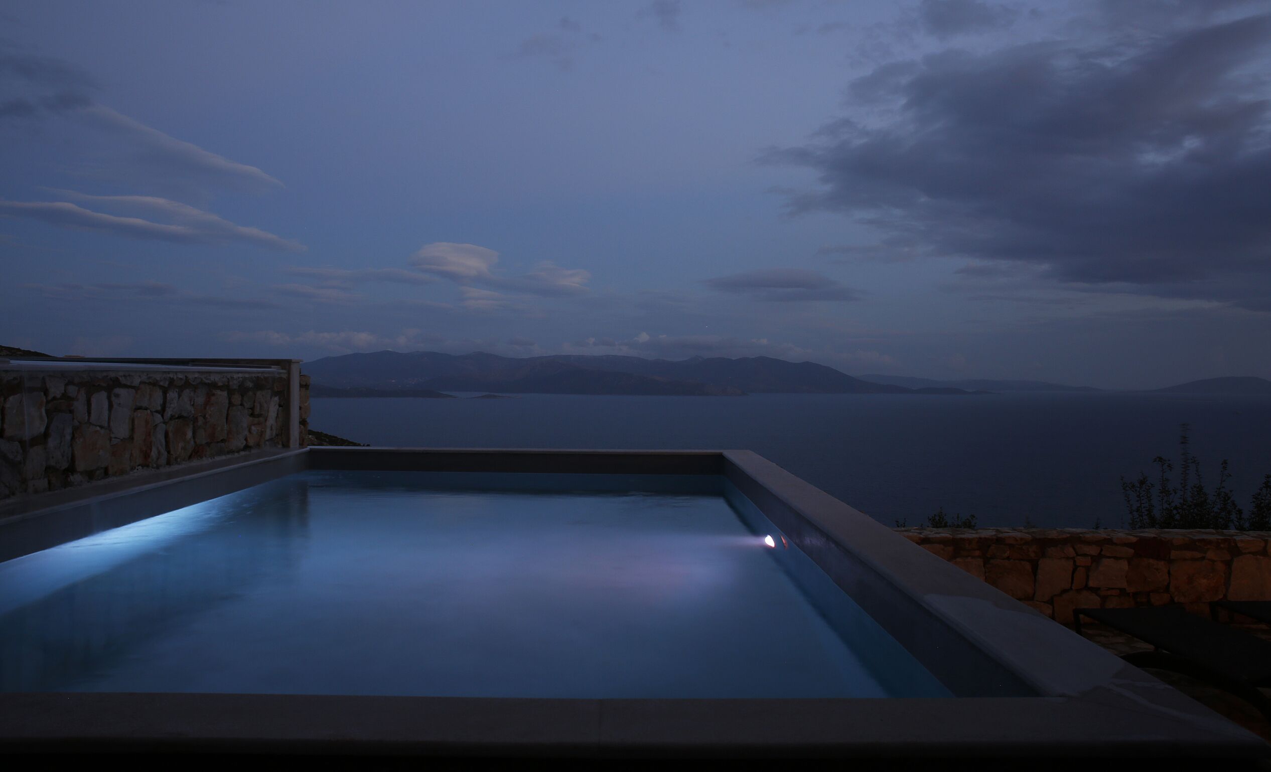 A swimming pool with a stone wall on one side and a view of the sea and mountains in the background.