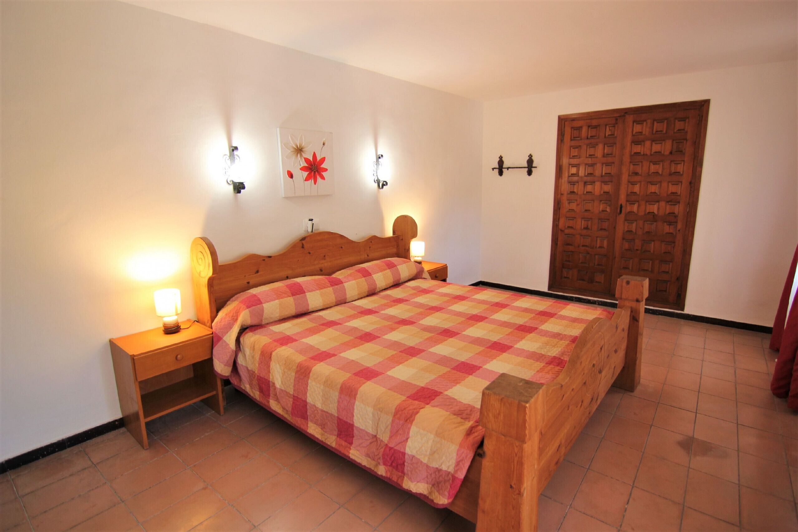 A bedroom with a wooden bed, a nightstand with a lamp, a wall-mounted light, and a wooden door.