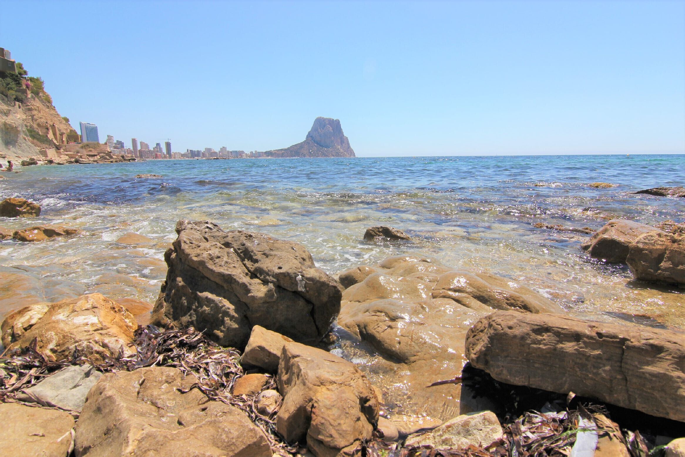 A rocky shoreline with clear water, a prominent rock formation in the distance, and a city skyline in the background.