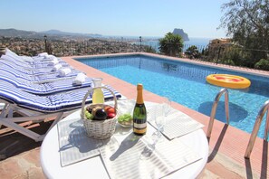 Un espace aménagé au bord de la piscine, avec des chaises longues, une table sur laquelle se trouvent une bouteille et des verres, et une vue sur la mer et le paysage urbain.