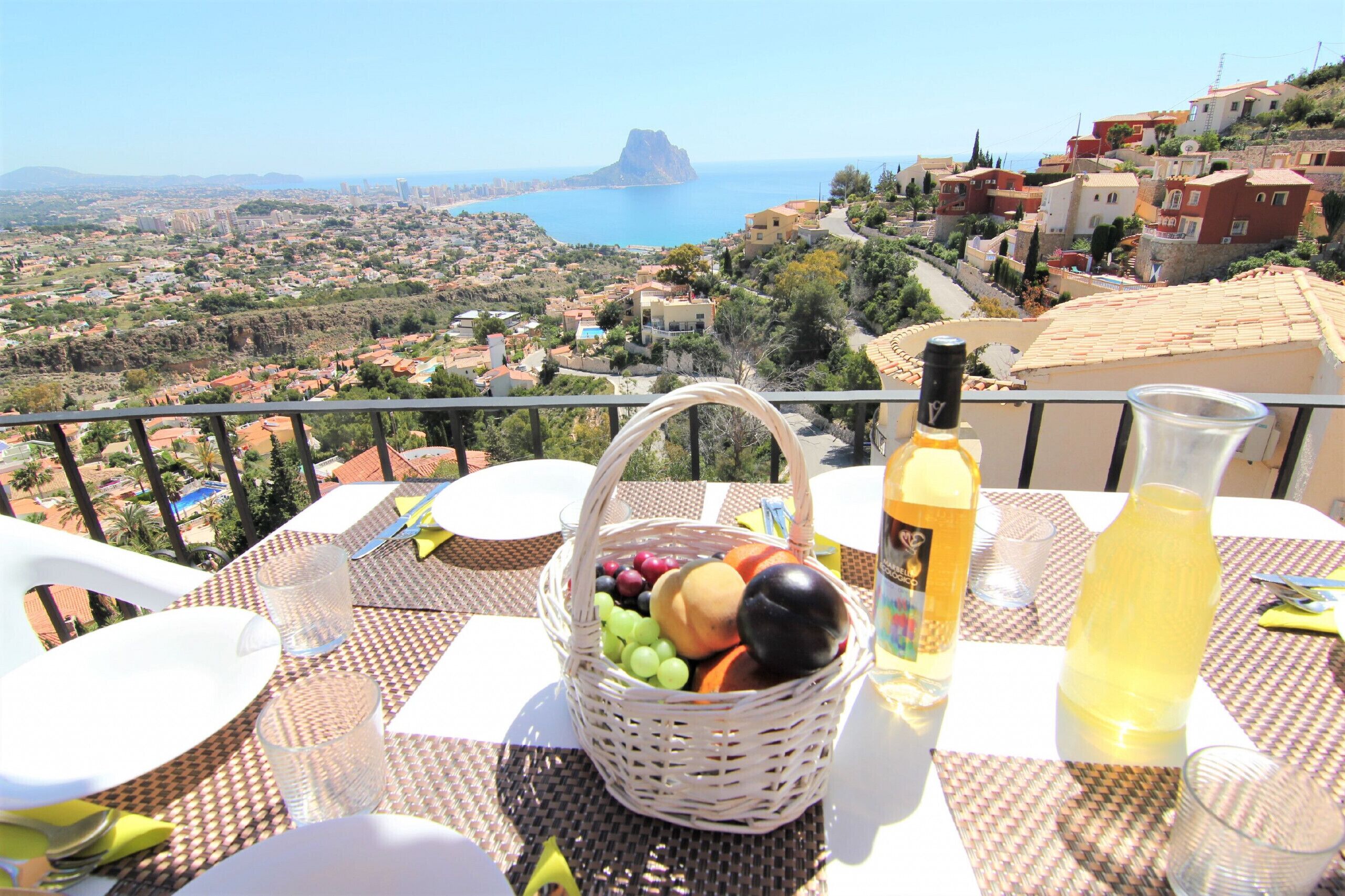 A table set with a fruit basket, a bottle, and a glass on a balcony overlooking a coastal town.