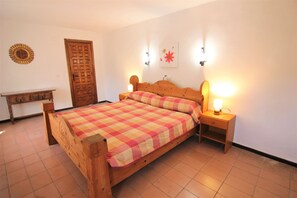 Un lit en bois recouvert d’une couverture à carreaux, une table de chevet avec une lampe, une porte en bois et une horloge murale.