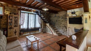 A rustic interior with a stone wall, wooden beams, a staircase, a television, and a kitchen area.