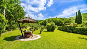 A well-maintained garden with a dining area featuring a table and chairs under a thatched umbrella, surrounded by lush greenery and a clear sky.