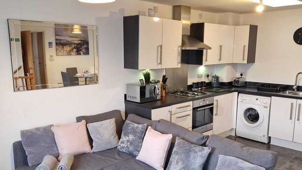 A modern living room with a grey sectional sofa, a glass coffee table, and a kitchen area with white cabinets and a washing machine. - HEREFORD – Robin Coach House: Modern 2-Bed with Parking on City Outskirts (Hereford)