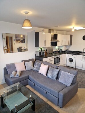 A modern living room with a grey sectional sofa, a glass coffee table, and a kitchen area with white cabinets and a washing machine.