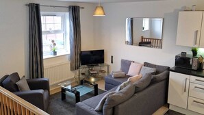 A modern living room with a grey sofa, a flat-screen TV, and a wooden staircase. - HEREFORD – Robin Coach House: Modern 2-Bed with Parking on City Outskirts (Hereford)