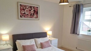 A neatly arranged bedroom with a bed, bedside tables, a lamp, a framed floral artwork, and a window with curtains. - HEREFORD – Robin Coach House: Modern 2-Bed with Parking on City Outskirts (Hereford)