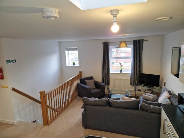 A living room with a grey sofa, a TV, a wooden staircase, and a fire exit sign.