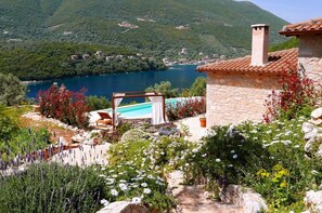 A stone villa with a pool, surrounded by lush greenery and a view of a lake and mountains.