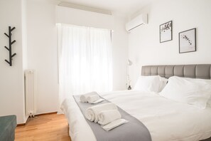 Een net opgemaakt bed met witte lakens, opgerolde handdoeken, een grijze plaid en een grijs hoofdbord.