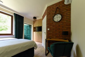 Un dormitorio con cama, escritorio, silla, televisor y pared de ladrillo con un reloj.