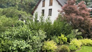 Ein weißes Haus mit geneigtem Dach, umgeben von dichtem Grün und verschiedenen Sträuchern.