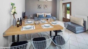 A dining area with a wooden table set for four, white wire chairs, a sofa, and a television in the background.