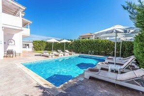 A swimming pool with lounge chairs and umbrellas, surrounded by greenery and buildings. - Villa Orchard - Four Bedroom Villa, Sleeps 9 (Zakynthos)
