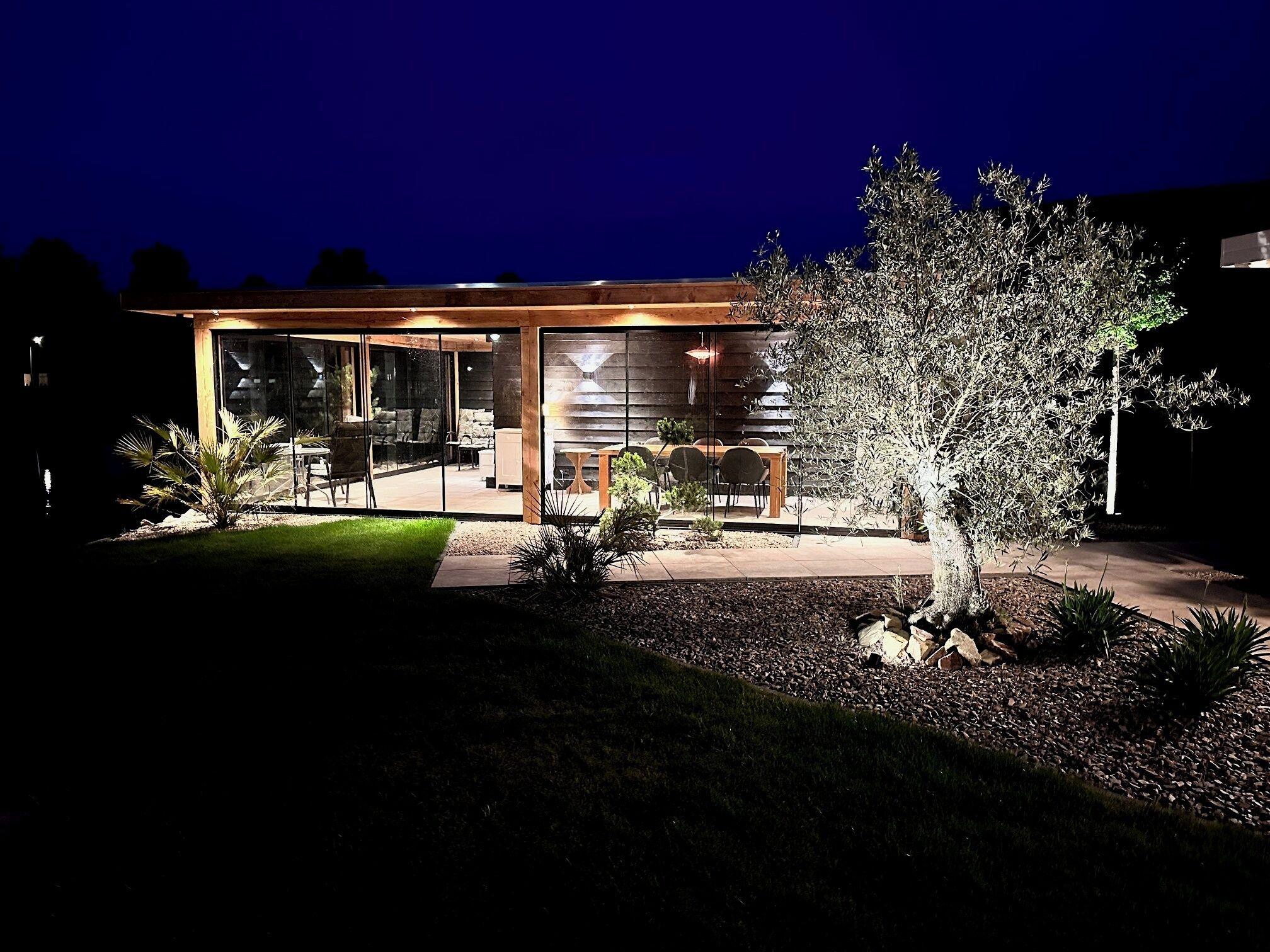 Ein modernes Haus mit Glasfassade, Sitzgelegenheit im Freien und einem gut beleuchteten Garten.