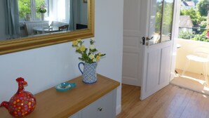 Une pièce comprenant une commode en bois, un vase avec des fleurs, une figurine de coq aux couleurs vives, un miroir et une porte donnant sur un balcon.