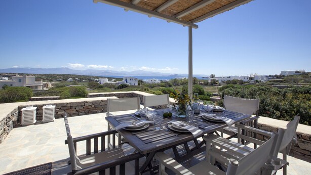 Un área de comedor al aire libre con una mesa preparada para cuatro, con vistas a un paisaje pintoresco de montañas y edificios.