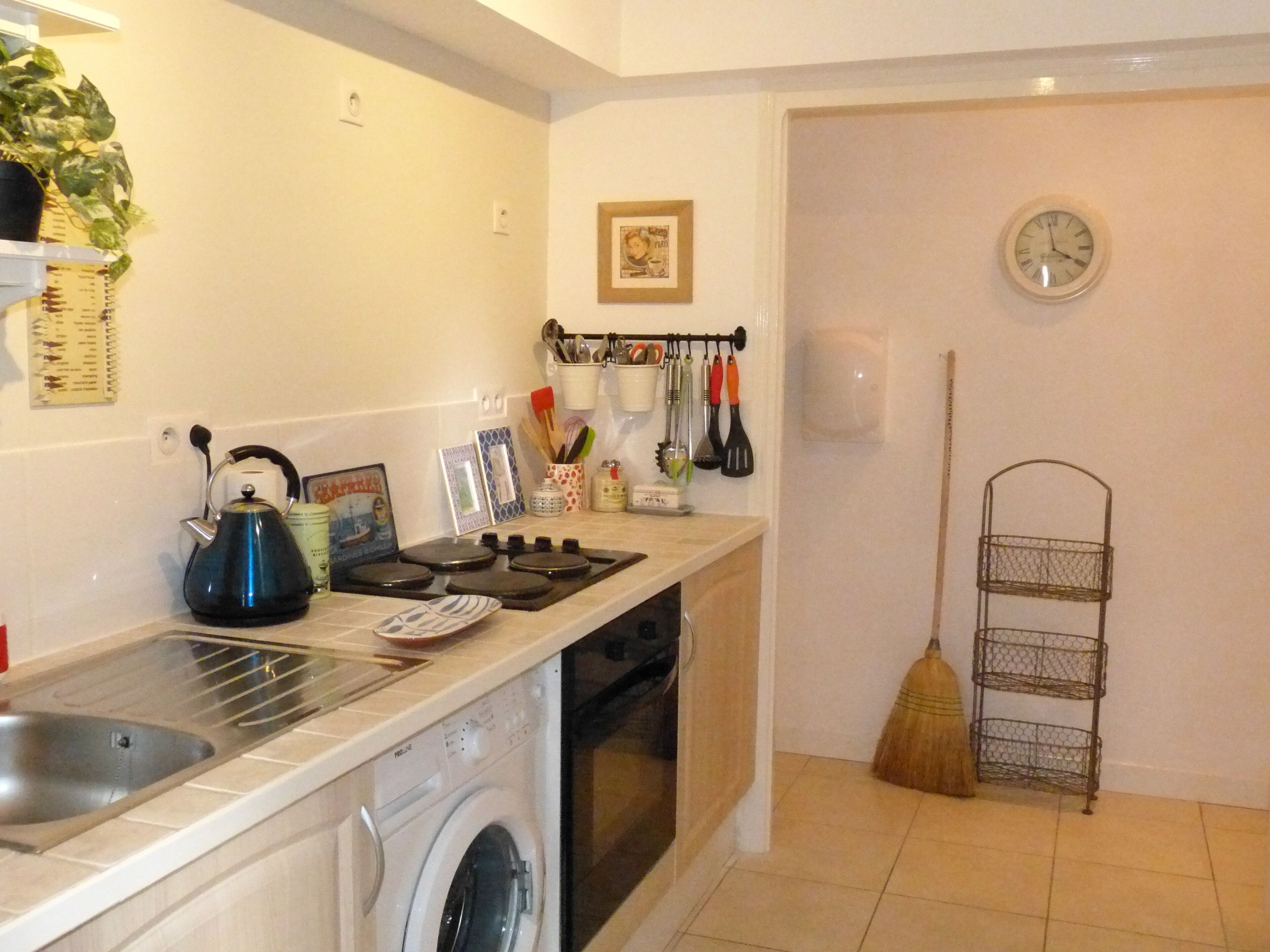 A compact kitchen with a washing machine, a kettle, and a sink. There is a broom and a metal rack on the floor.