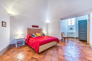 A bedroom with a large bed, a nightstand, a chair, and a door leading to another room.