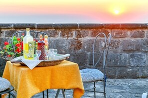 Ein Tisch, gedeckt mit Flaschen, Gläsern und einer Schale, mit einem Stuhl, im Hintergrund ein Sonnenuntergang.