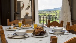 A dining table with plates of food, a glass of orange juice, and a view of a green landscape through a balcony door. - SAMAREITIDOS LIGHT STUDIO  - GREEN (Anafotia)
