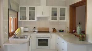 Una cucina compatta con mobili bianchi, forno a incasso, lavello e un piccolo angolo con bollitore, frullatore e una ciotola di frutta.