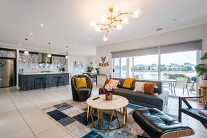 A modern living room with a sofa, a coffee table, and a dining area with a kitchen in the background. - Waterfront Grand Villa in Sanctuary Lakes (Point Cook)