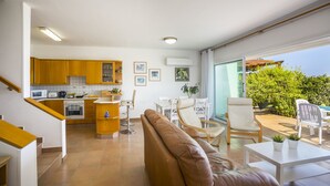 A modern kitchen with wooden cabinets, a dining area with a white table and chairs, and a large window overlooking a garden. - Sirina Lina Beachfront Suite (Protaras)