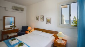 A hotel room with a bed, bedside tables, a desk, a chair, a window with a view, and wall-mounted pictures. - Sirina Lina Beachfront Suite (Protaras)