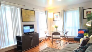 Ein Wohnzimmer mit Sofa, Fernseher auf einem Schrank, einem Esstisch mit Stühlen und einer Pflanze.