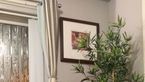 Ein Eckzimmer mit blauem Sessel, einer Topfpflanze, einem Fenster mit Vorhängen und einem gerahmten Bild an der Wand.