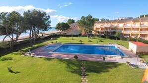 A swimming pool surrounded by a well-maintained lawn, with a beach and hotel buildings in the background.