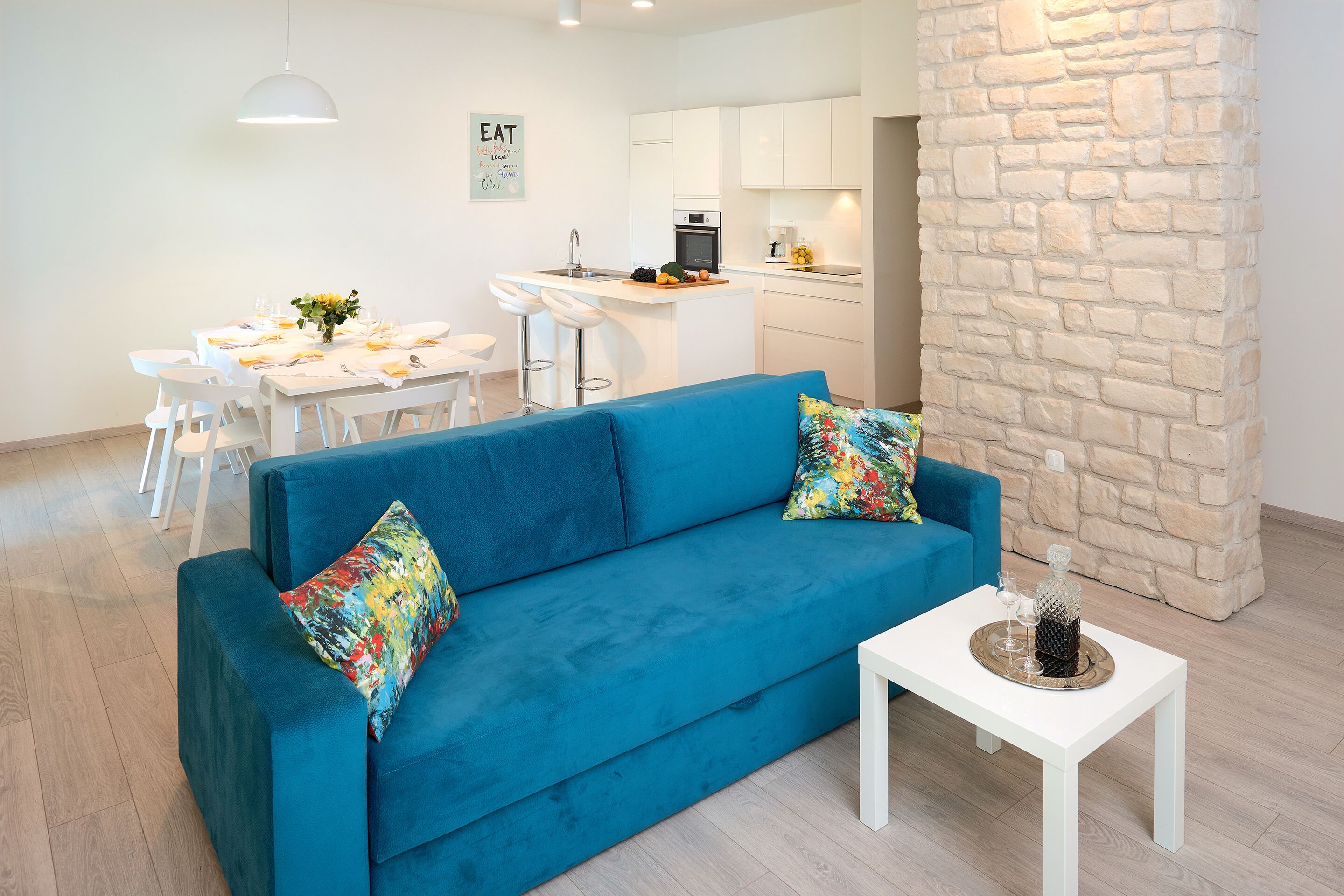 Een moderne woonkamer met een blauwe bank, een witte salontafel en een keuken op de achtergrond.