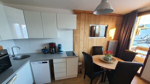 A compact kitchen with white cabinets, a coffee maker, a kettle, and a dishwasher. There is a dining area with a table, chairs, and a vase of flowers. A large window with curtains is visible, offering a view of a snowy landscape. - Studio with mountain corner 1 (AVORIAZ)