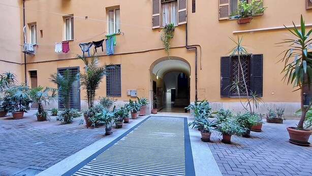 An outdoor courtyard with a central pathway, potted plants, and a building with multiple windows and balconies. - Red House Rome - Trastevere (Roma)