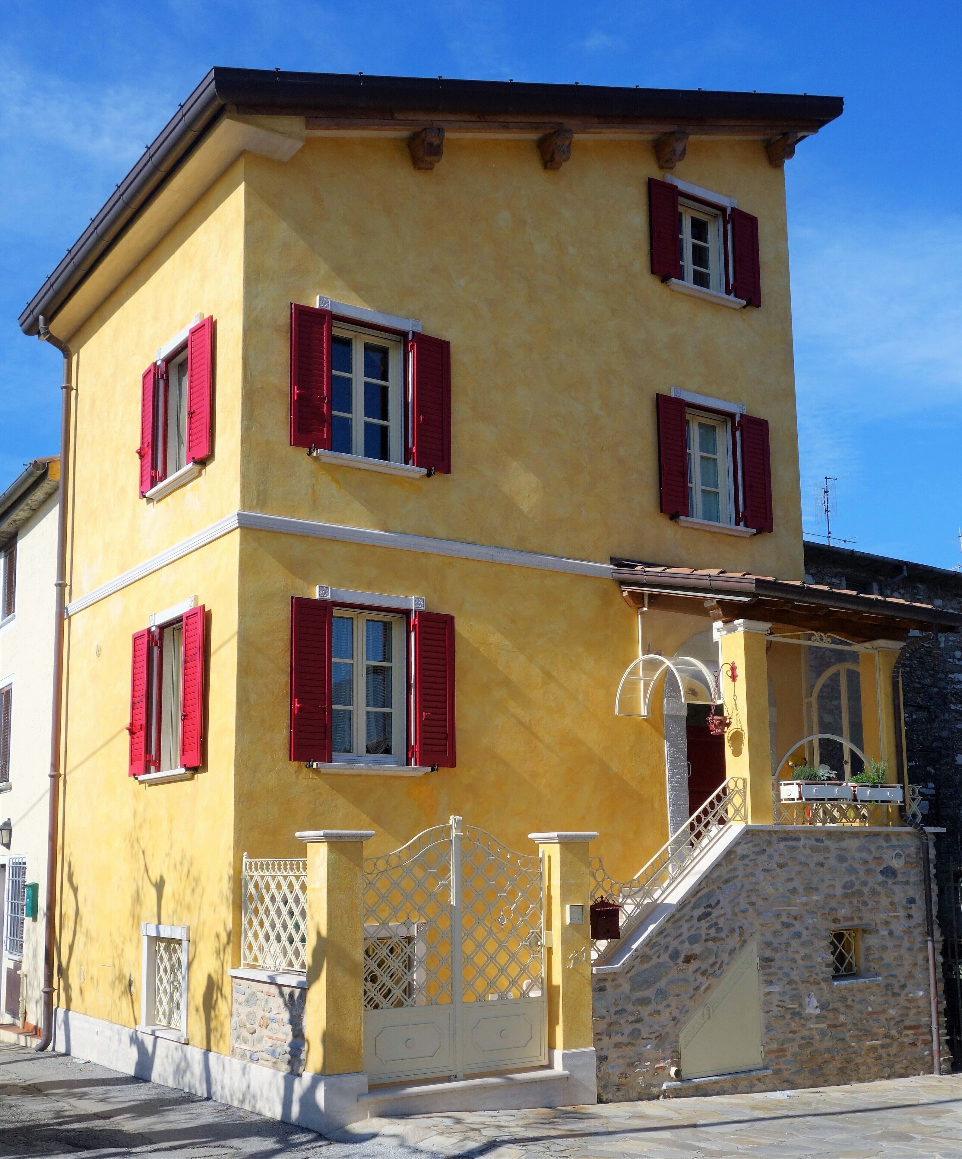 Un immeuble de deux étages avec des volets rouges, un escalier en pierre et un portail en métal.