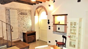 Ein gemütliches Zimmer mit Steinwand, Holzbalken, Kamin, Esstisch mit Buch und einem Sessel.