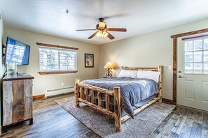 A bedroom with a wooden bed, a ceiling fan, a nightstand with a lamp, a TV on a wooden cabinet, and a window with a view of buildings. - Modern Mt Retreat |1 Mile from Steamboat Resort! (Steamboat Springs)