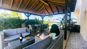 A covered outdoor seating area with wicker furniture, a table with a bottle of wine, and a glass of red wine. - Hann's Villa - Charming Kapparis Getaway (Protaras)