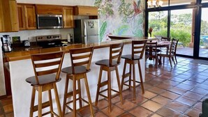 A kitchen with wooden cabinets, a microwave, a stove, and a refrigerator. There are three wooden bar stools and a dining table with chairs.