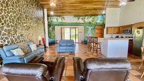 A living room with a stone wall, a blue sofa, a blue armchair, and a leather ottoman. There is a kitchen area with wooden cabinets and a refrigerator.