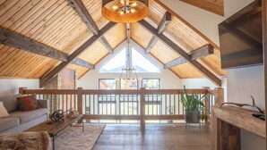 A spacious loft with a high ceiling, wooden beams, a large window, and a cozy seating area with a sofa and a coffee table. - Blissful Mountain Getaway – Steps to Lake Estes, Indoor/Outdoor Fireplace (Estes Park)