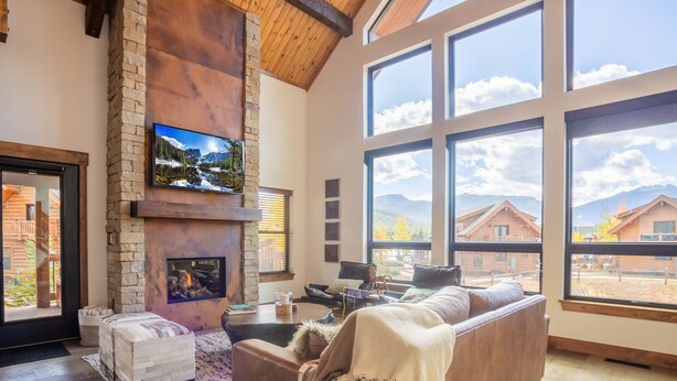 A spacious living room with a stone fireplace, a large TV mounted on the wall, and a view of the mountains through large windows. - Blissful Mountain Getaway – Steps to Lake Estes, Indoor/Outdoor Fireplace (Estes Park)