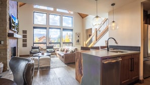 A spacious living area with a fireplace, a large window overlooking a scenic view, a kitchen with a modern island, and a wooden staircase. - Blissful Mountain Getaway – Steps to Lake Estes, Indoor/Outdoor Fireplace (Estes Park)