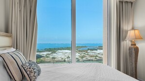 Ein Hotelzimmer mit einem großen Bett, beigen Vorhängen, Blick auf Strand und Meer und einer Lampe am Rand.