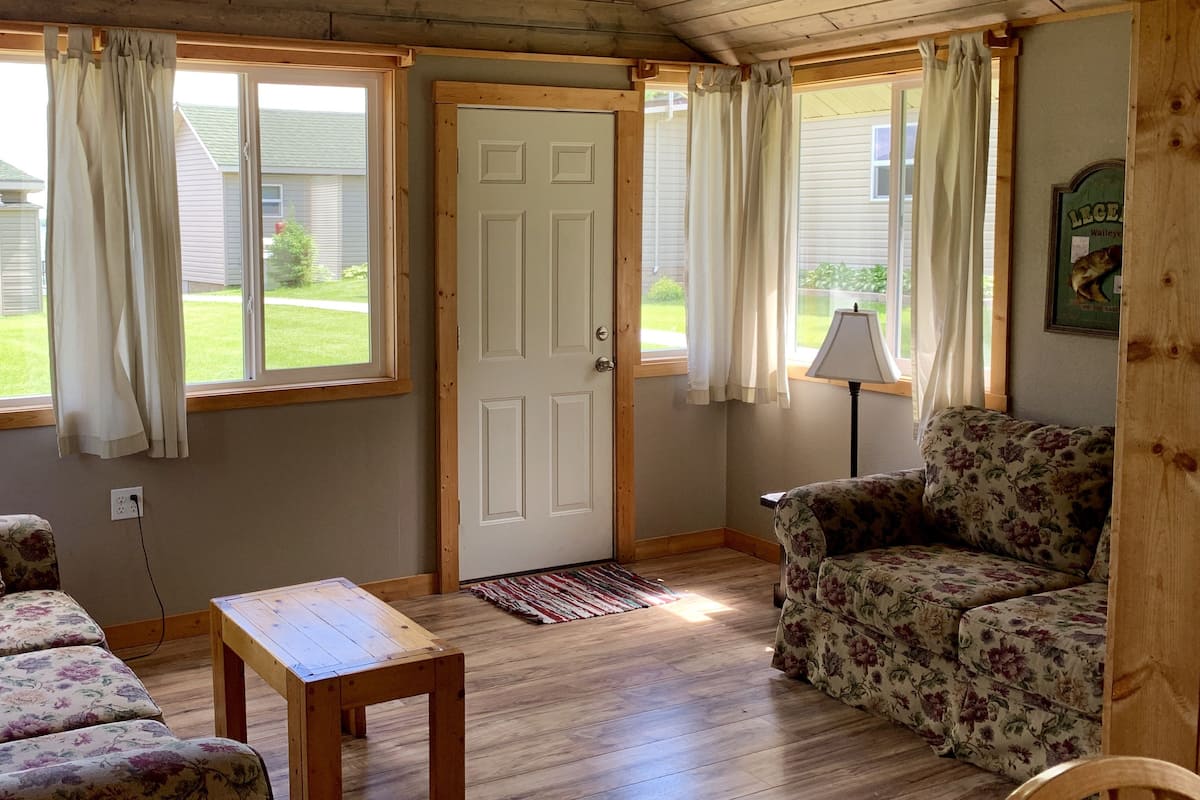 A cozy living room with wooden flooring, a floral sofa, a wooden coffee table, and a door leading outside.